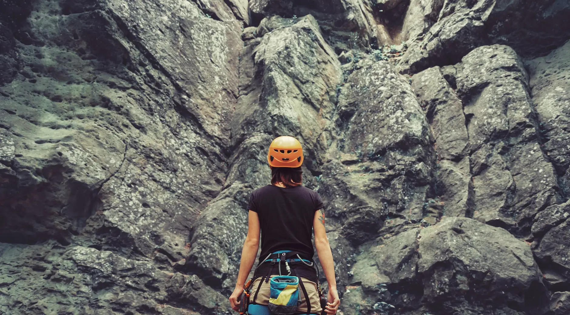 Rock climber in helmet and harness facing tall stone cliff face